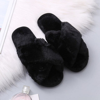 Black fluffy slippers on a light gray surface with a bottle of perfume and fur rug.
