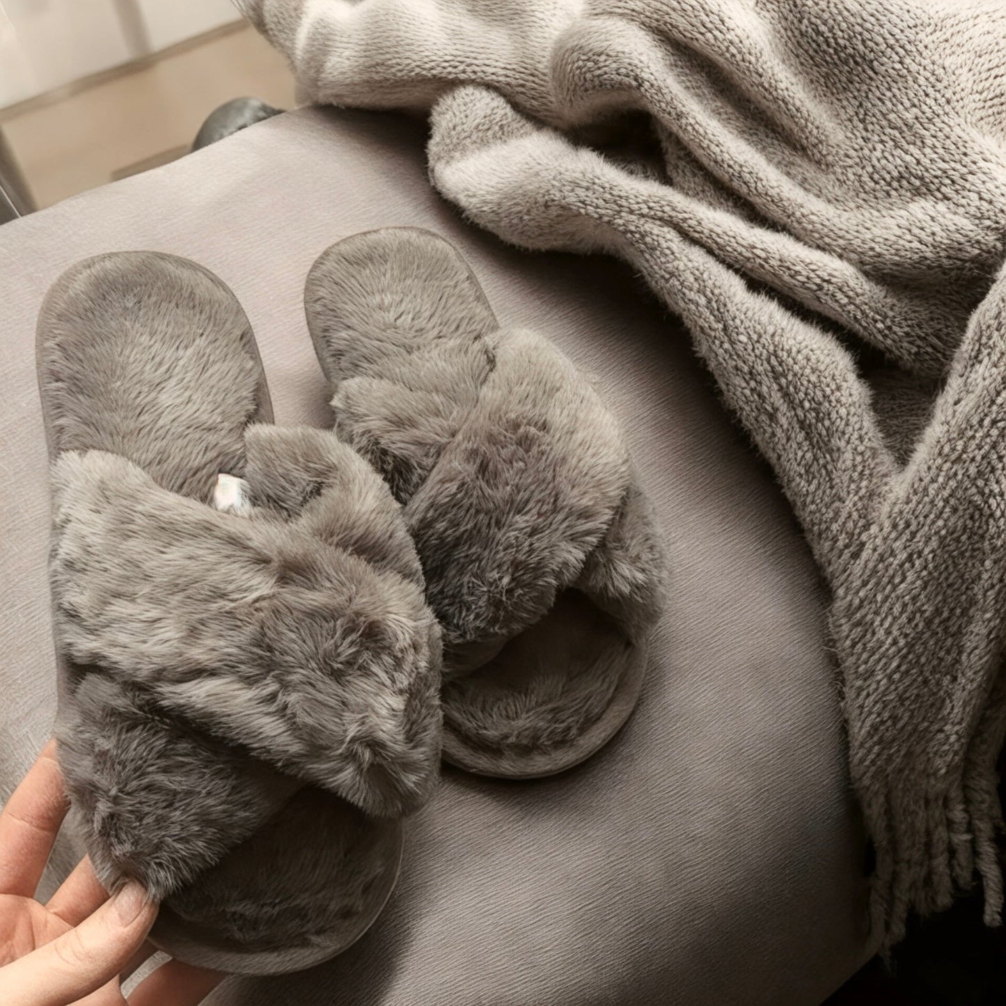 Gray fuzzy slippers on a cushion with a blanket in the background