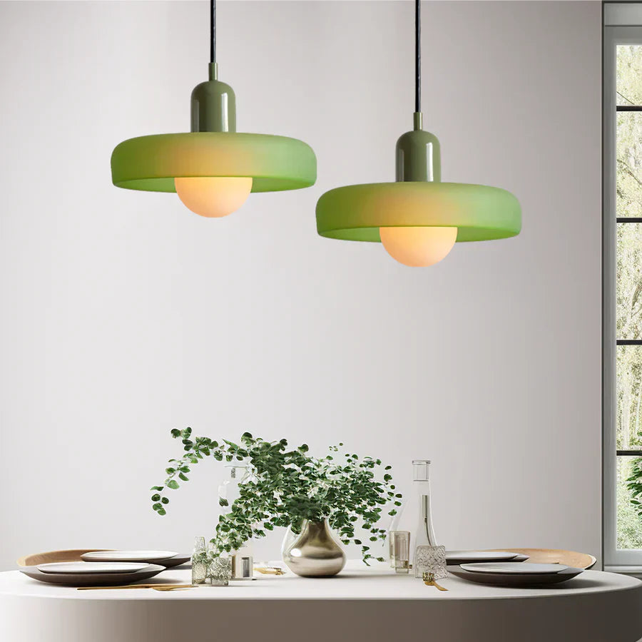 Two green pendant lights hanging above a dining table with a plant and glasses.