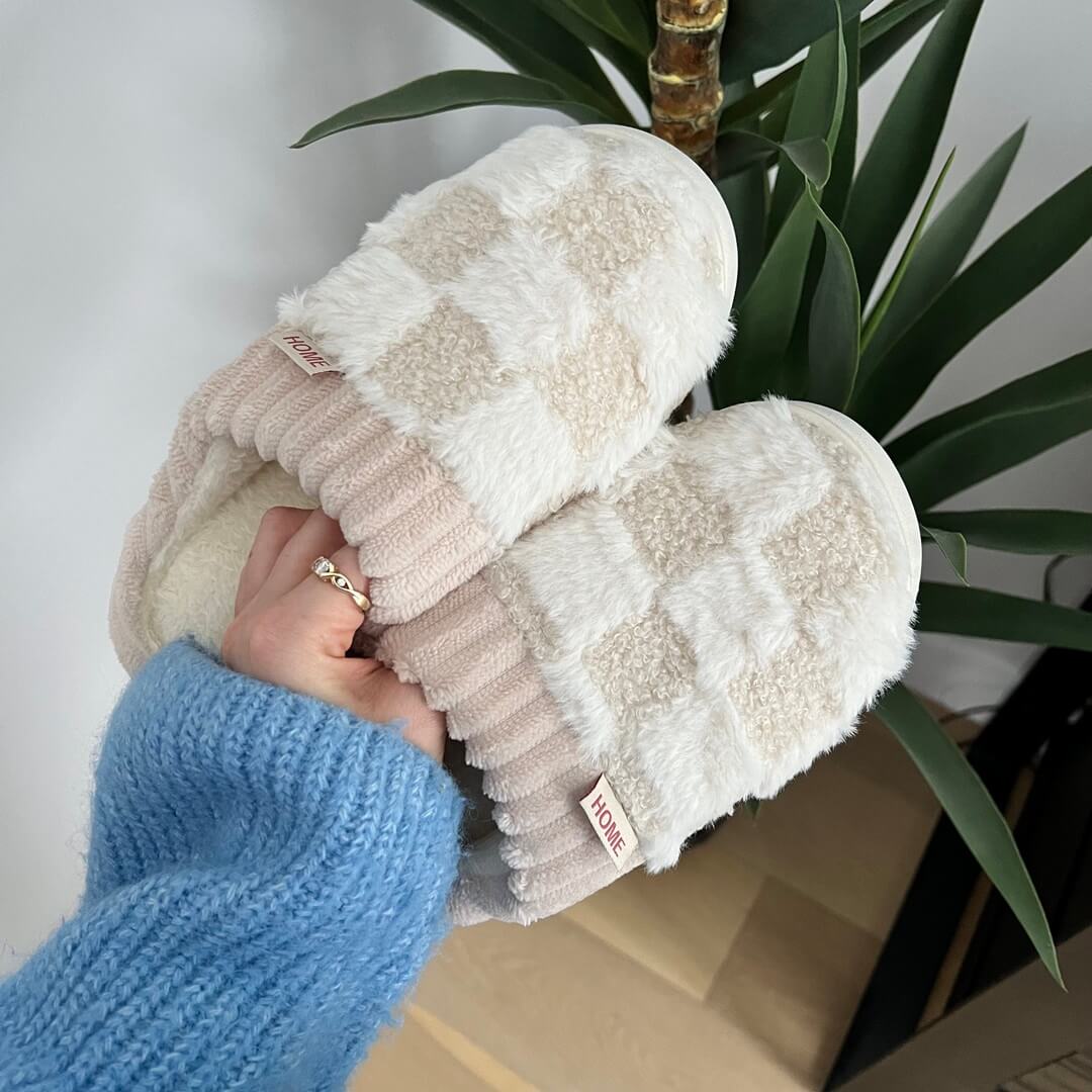 Pair of fluffy slippers held by a person wearing a blue sweater, with a plant in the background.
