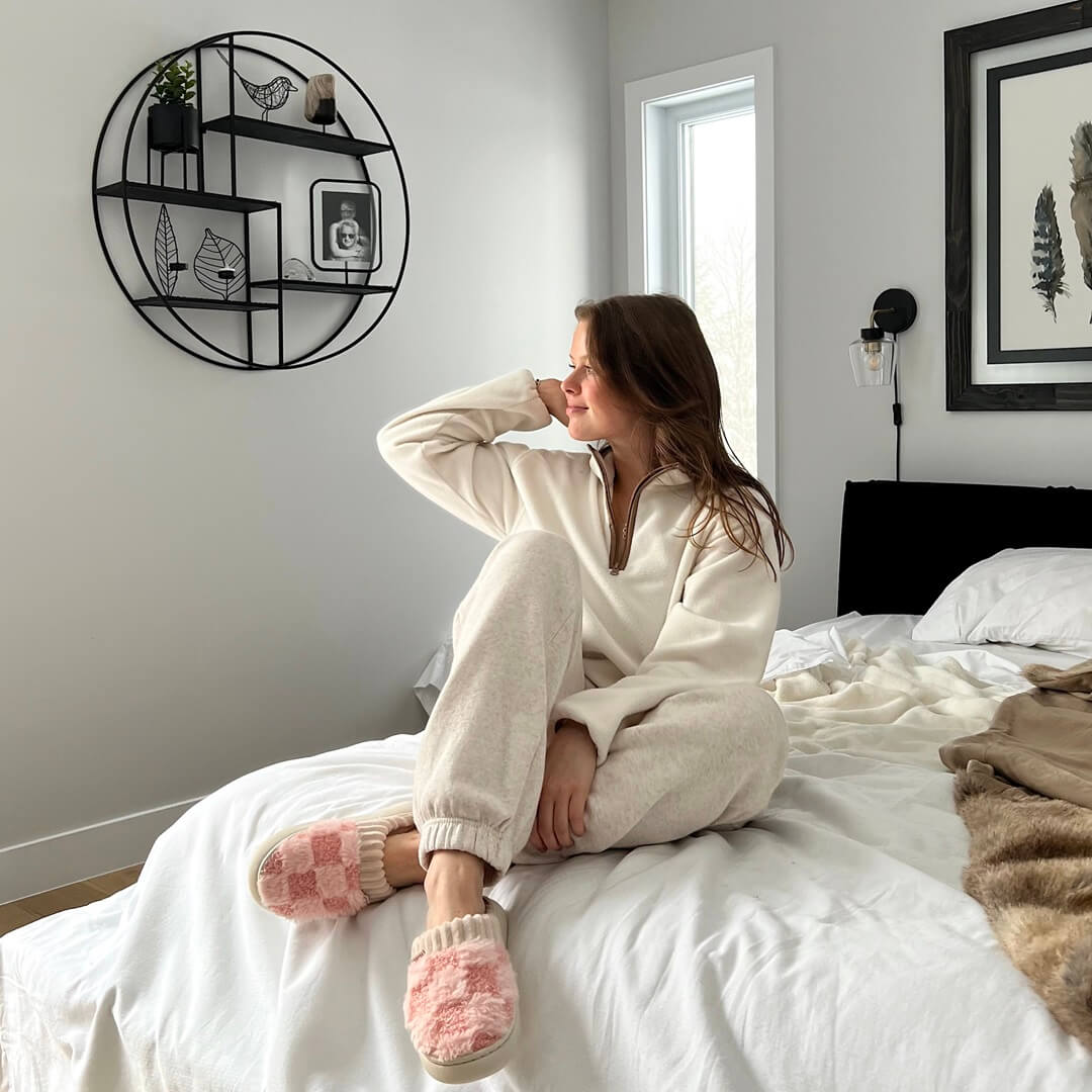 Woman in a cozy room sitting on a bed wearing a beige outfit and pink slippers.