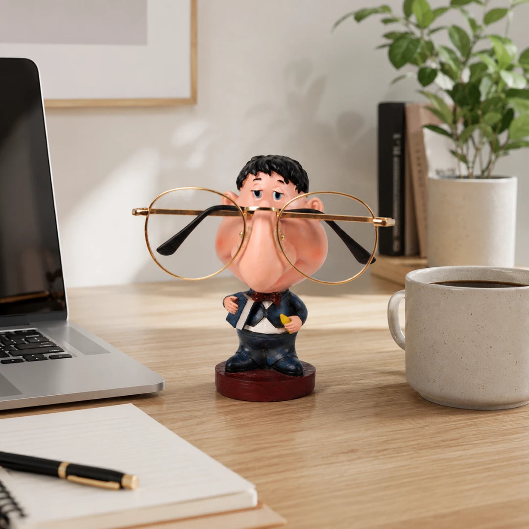 Josh & Tate - NoseNook™ Glasses Figurine Stand - A novelty desk accessory featuring a cartoon character wearing round gold-framed glasses, styled on a wooden desk next to a laptop, notebook, coffee mug, and potted plant.