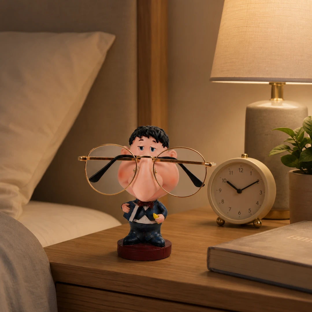 Josh & Tate - NoseNook™ Glasses Figurine Stand - A novelty glasses holder featuring a cartoon man in a suit holding oversized gold-rimmed glasses, displayed on a wooden bedside table next to a lamp, alarm clock, and book in a warm bedroom setting.