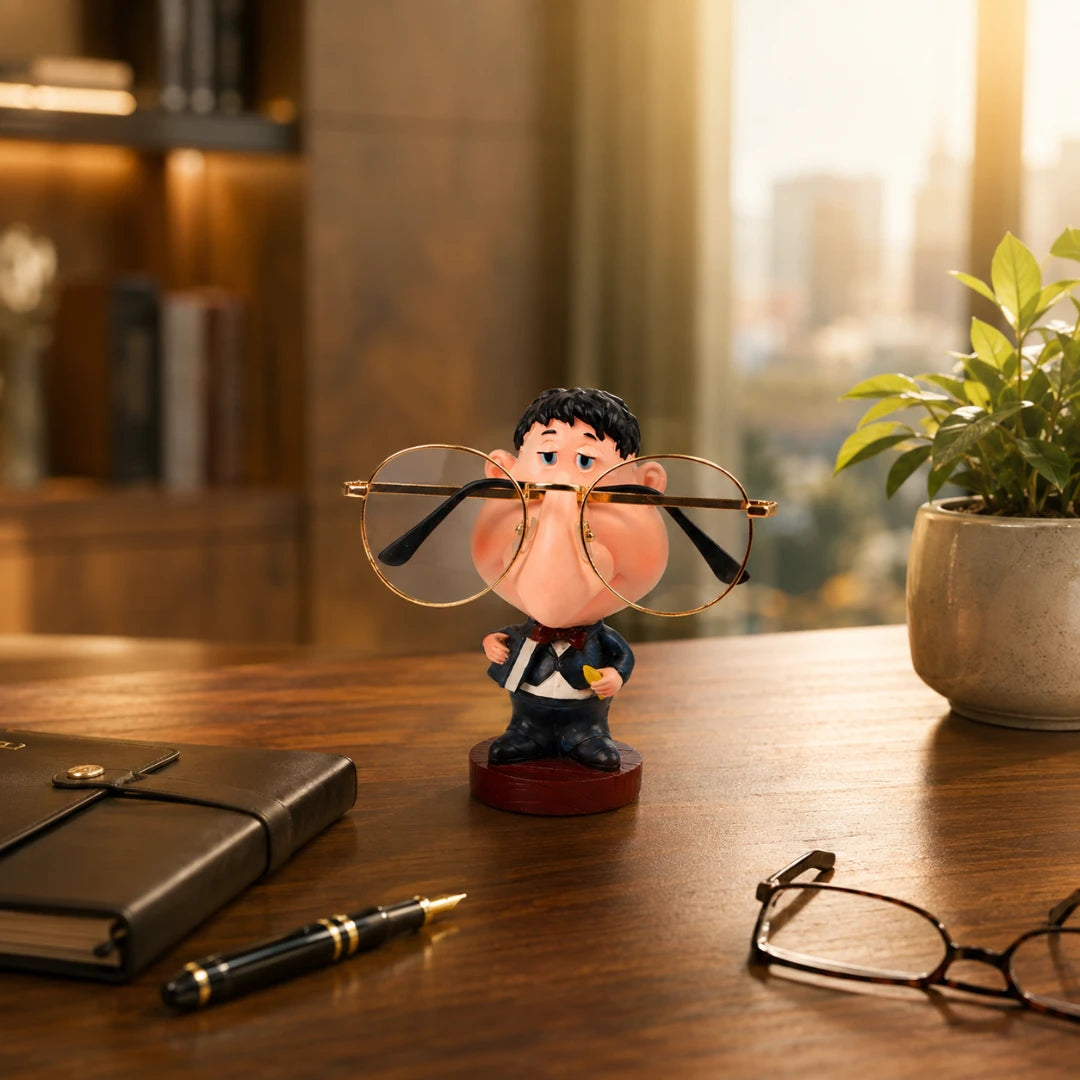 Josh & Tate - NoseNook™ Glasses Figurine Stand - A novelty desk figurine of a cartoon character wearing round gold-rimmed eyeglasses, styled on a wooden desk with a notebook, pen, and another pair of glasses in a warm, blurred office setting.