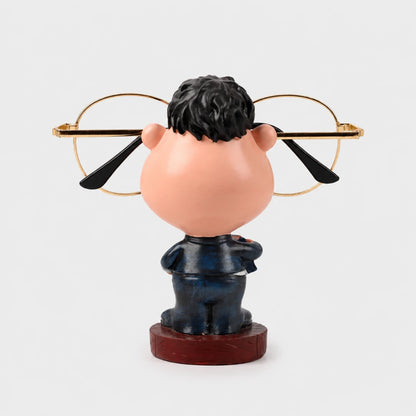 Josh & Tate - NoseNook™ Glasses Figurine Stand - A novelty bobblehead figurine of a man with a large peach-colored head, black hair, and a dark blue suit standing on a round wooden base, holding gold-framed eyeglasses behind his head.