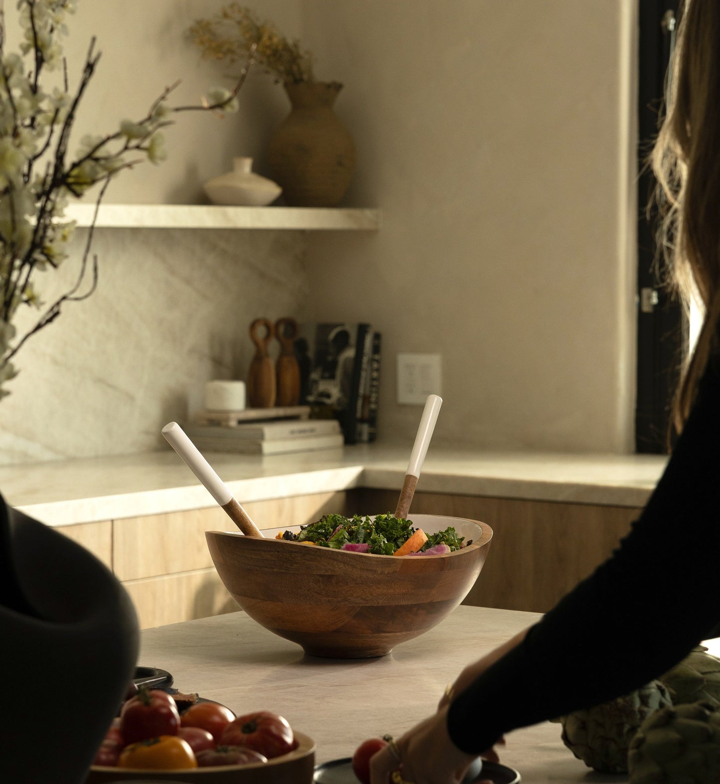 Person preparing food in a kitchen with a wooden bowl of salad on a counter.