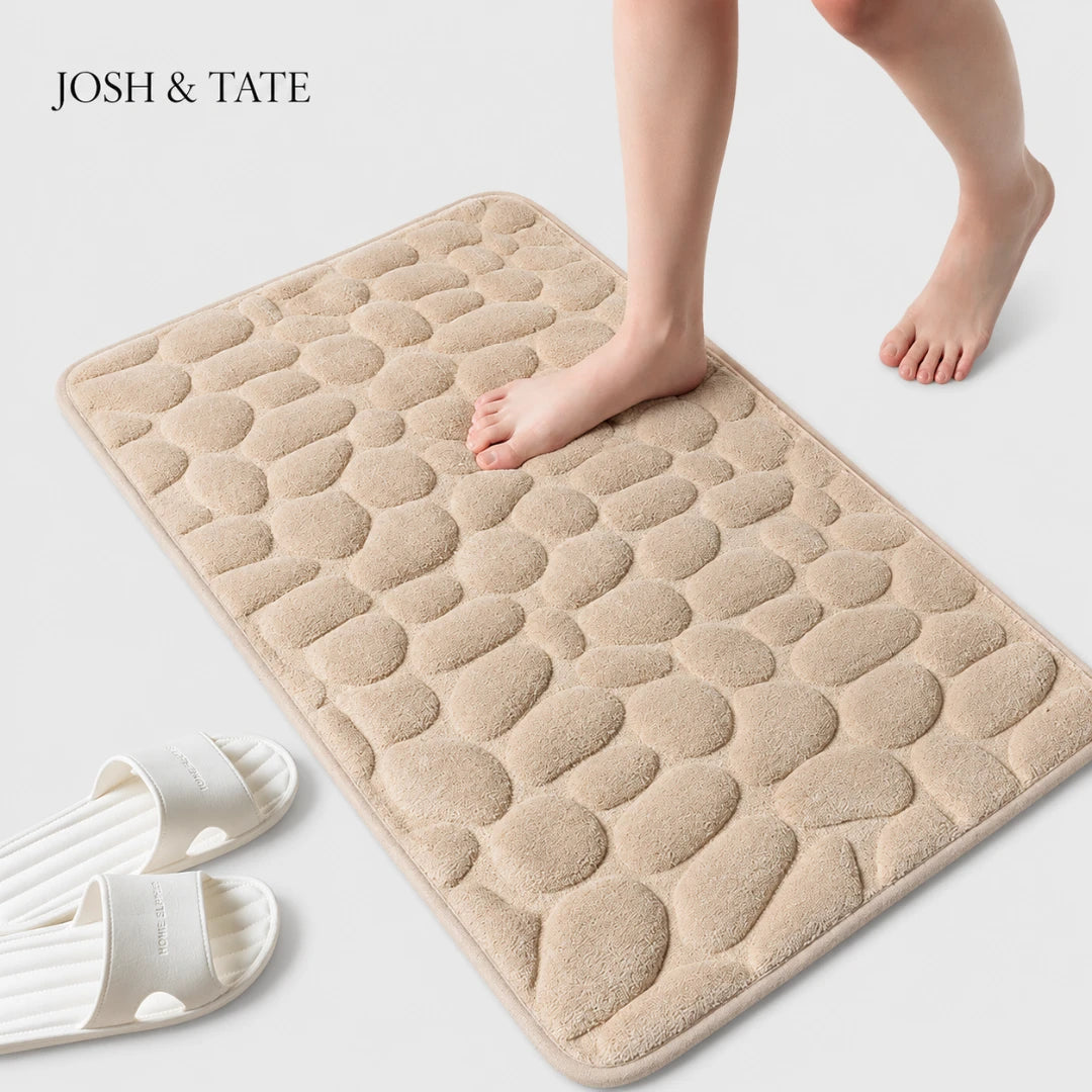 Josh & Tate - PebbleSoft™ Non-Slip Memory Foam Mat - Beige textured bath mat with feet stepping onto it, next to white slippers on a light gray background.