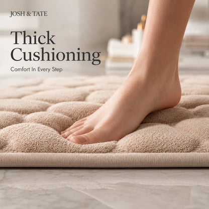 Josh & Tate - PebbleSoft™ Non-Slip Memory Foam Mat - Beige bath mat with thick cushioning, foot stepping onto it, and 'Josh & Tate' branding.