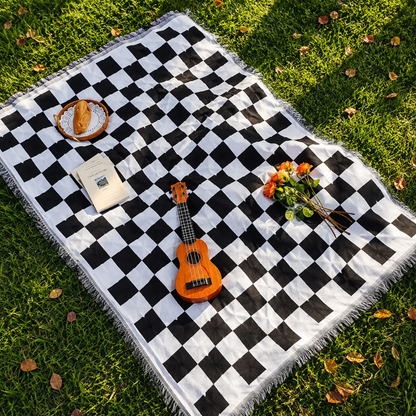 Josh & Tate - PicnicCo™ Woven Outdoor Blanket - Checkered picnic blanket with a small guitar, flowers, and a pastry on a grassy field.