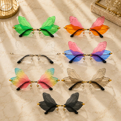 Josh & Tate - PixieFrame™ Butterfly Sunglasses - Seven pairs of butterfly-wing style sunglasses arranged on a light marble surface. The translucent wing frames come in green, orange, blue, pink-to-yellow gradient, smoky beige, and dark gray/black, with gold metal temples and black temple tips.