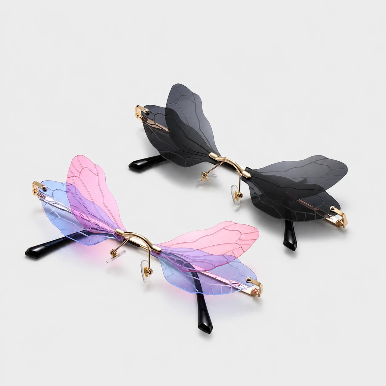 Josh & Tate - PixieFrame™ Butterfly Sunglasses - Two pairs of butterfly-wing style eyeglasses on a light gray background. One pair has translucent dark gray wings with gold metal arms and black temple tips.