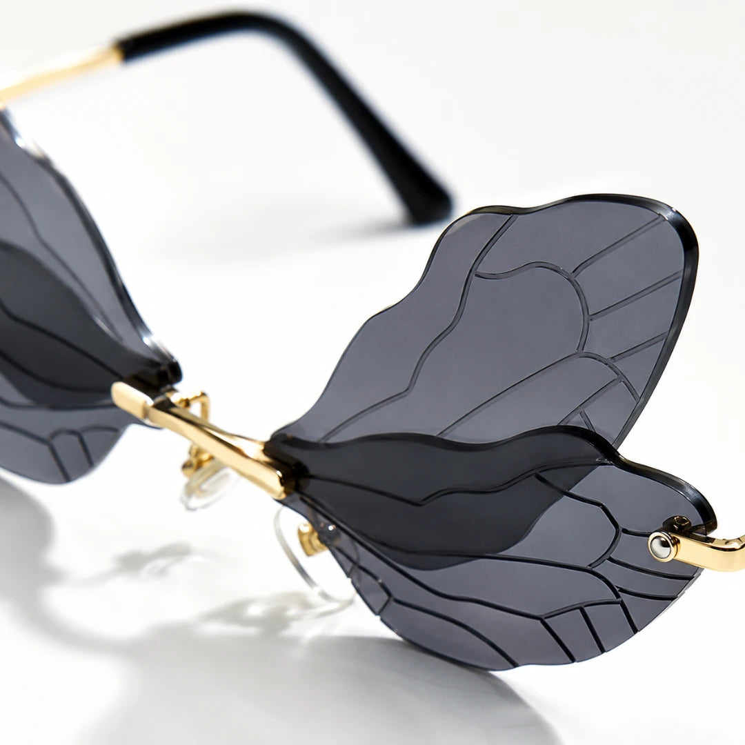 Josh & Tate - PixieFrame™ Butterfly Sunglasses - Close-up of a pair of rimless butterfly-style sunglasses with dark gray tinted lenses, gold metal hinges, and black temple arms on a white background.
