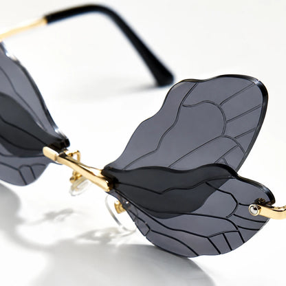 Josh & Tate - PixieFrame™ Butterfly Sunglasses - Close-up of a pair of rimless butterfly-style sunglasses with dark gray tinted lenses, gold metal hinges, and black temple arms on a white background.