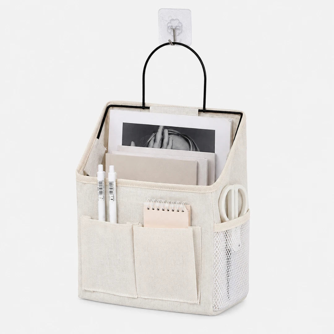 Josh & Tate - PocketPerch™ Hanging Organizer - A beige fabric desk organizer hanging from a clear wall hook, featuring a black metal handle, front pockets, a side mesh pocket, and stationery items inside on a white background.