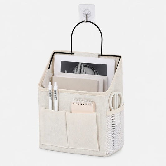 Josh & Tate - PocketPerch™ Hanging Organizer - A beige fabric desk organizer hanging from a clear wall hook, featuring a black metal handle, front pockets, a side mesh pocket, and stationery items inside on a white background.