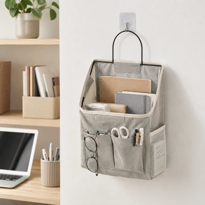 Josh & Tate - PocketPerch™ Hanging Organizer - A light gray fabric hanging organizer pouch with a black metal handle, mounted on a white wall with a clear hook. The organizer contains notebooks, glasses, scissors, and pens, with a blurred home office shelf scene on the left.