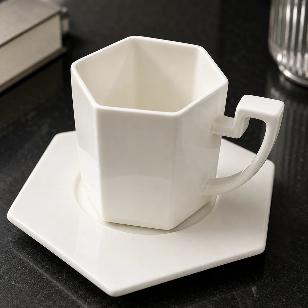 Josh & Tate - PrismCup™ Hexagon Cup & Saucer - A white hexagonal ceramic mug with a matching square saucer, set on a dark tabletop with a blurred book and glass in the background.