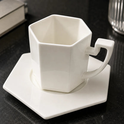 Josh & Tate - PrismCup™ Hexagon Cup & Saucer - A white hexagonal ceramic mug with a matching square saucer, set on a dark tabletop with a blurred book and glass in the background.