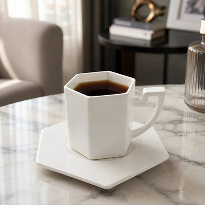 Josh & Tate - PrismCup™ Hexagon Cup & Saucer - A white hexagonal ceramic mug filled with black coffee sits on a matching white saucer on a marble table, with a softly blurred living room background.