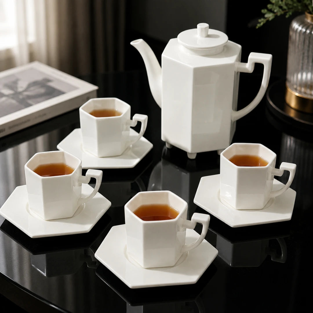 Josh & Tate - PrismCup™ Hexagon Cup & Saucer - A white ceramic tea set with a hexagonal teapot, four matching cups on saucers, and tea, arranged on a glossy black table in an indoor setting.