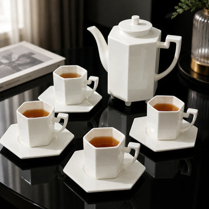 Josh & Tate - PrismCup™ Hexagon Cup & Saucer - A white ceramic tea set with a hexagonal teapot, four matching cups on saucers, and tea, arranged on a glossy black table in an indoor setting.