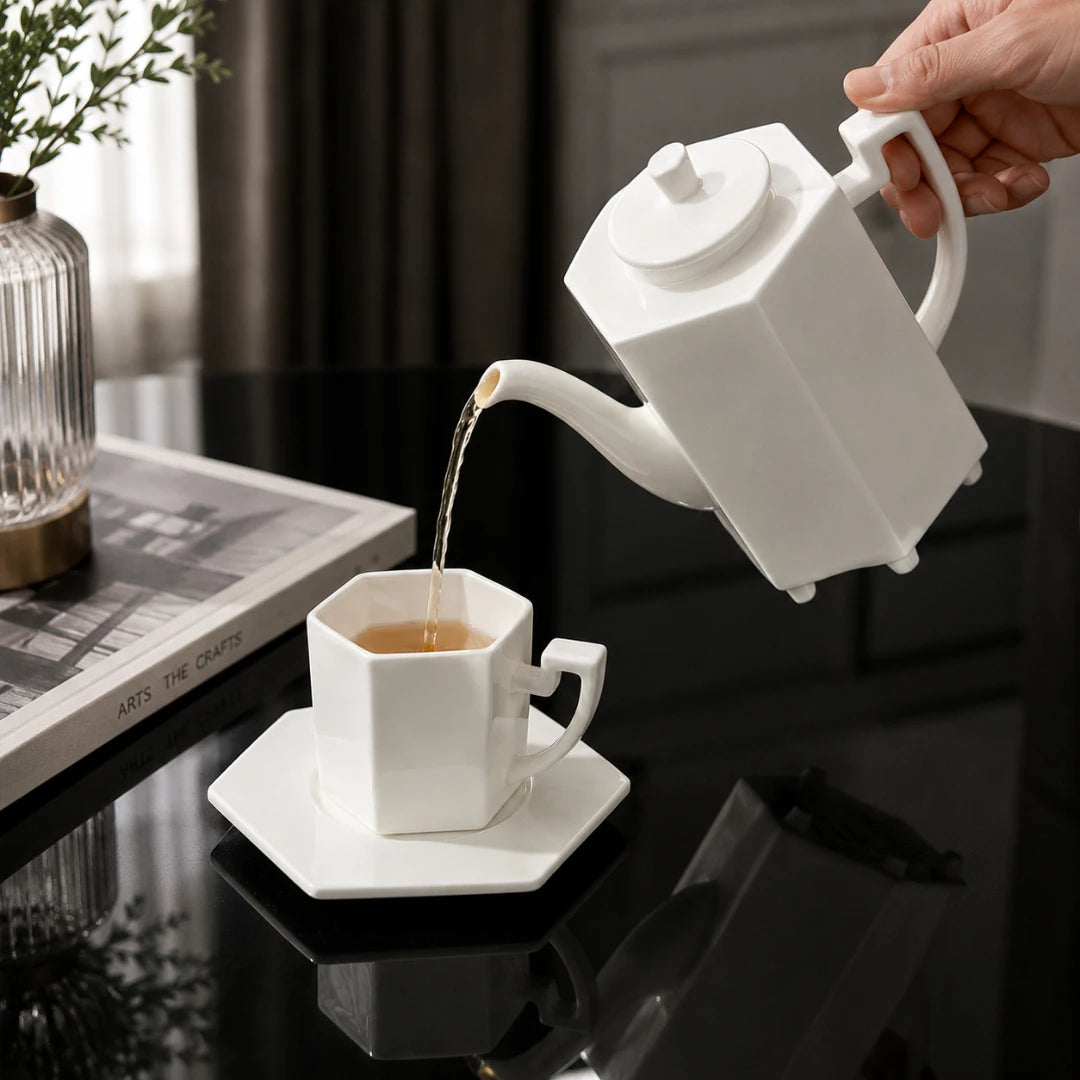 Josh & Tate - PrismCup™ Hexagon Cup & Saucer - A white ceramic teapot with a long spout is pouring tea into a matching white hexagonal teacup and saucer set on a glossy black table. A hand holds the teapot, and a book and vase are visible in the background.