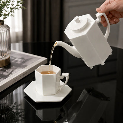 Josh & Tate - PrismCup™ Hexagon Cup & Saucer - A white ceramic teapot with a long spout is pouring tea into a matching white hexagonal teacup and saucer set on a glossy black table. A hand holds the teapot, and a book and vase are visible in the background.