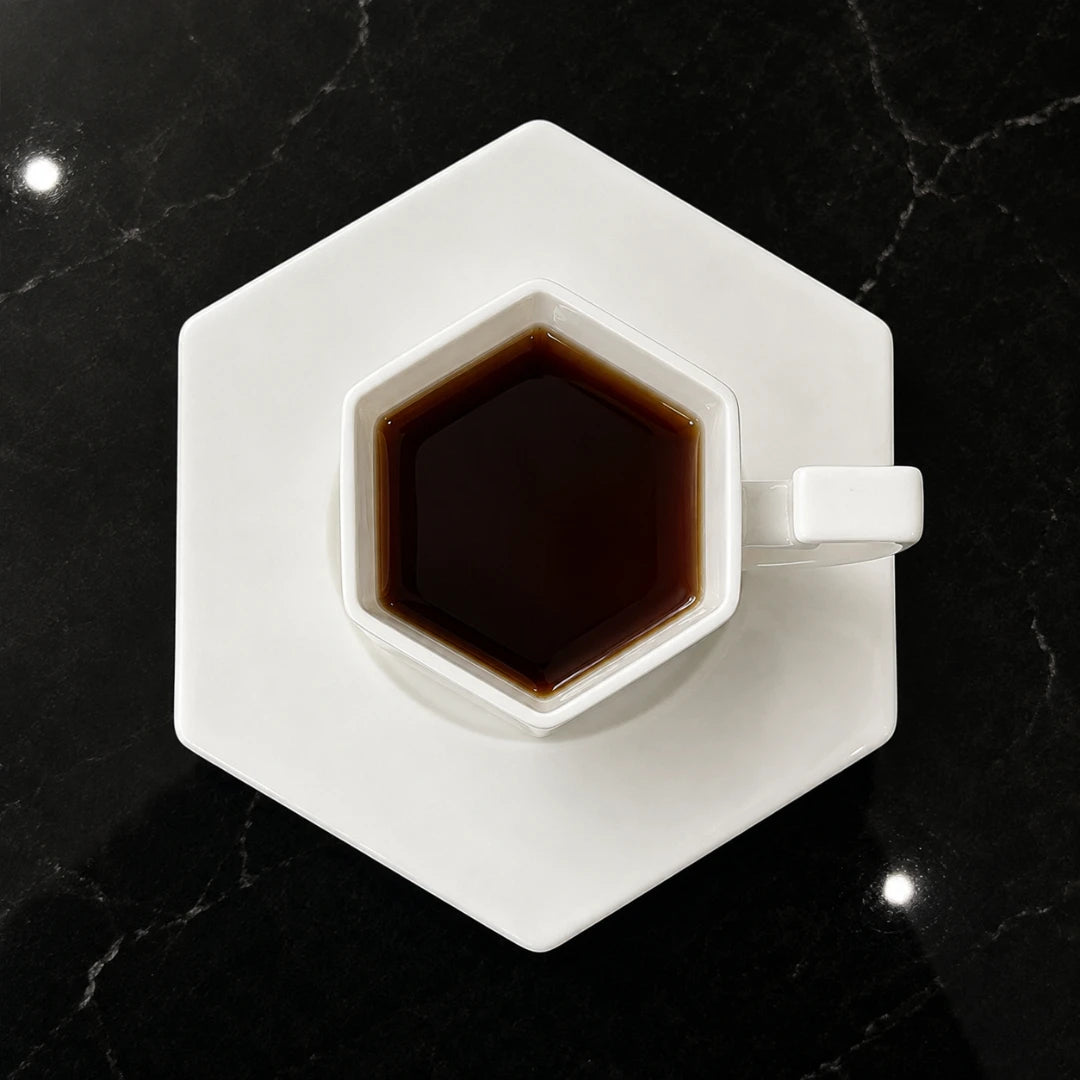 Josh & Tate - PrismCup™ Hexagon Cup & Saucer - A hexagonal white ceramic coffee cup and matching saucer, filled with dark coffee, placed on a black marble surface.