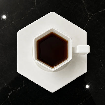 Josh & Tate - PrismCup™ Hexagon Cup & Saucer - A hexagonal white ceramic coffee cup and matching saucer, filled with dark coffee, placed on a black marble surface.
