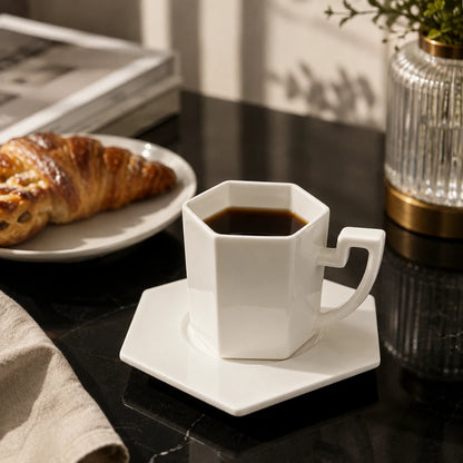 Josh & Tate - PrismCup™ Hexagon Cup & Saucer - A white hexagonal ceramic coffee cup filled with black coffee sits on a matching white saucer on a dark tabletop. A croissant is on a plate in the background, with a glass vase and sunlight casting shadows around it.