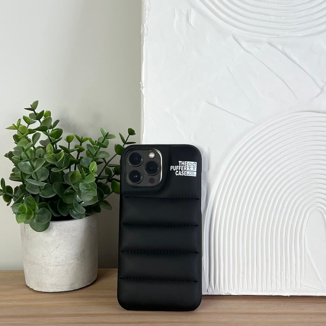 Black puffer phone case on a wooden surface with a plant and textured wall in the background