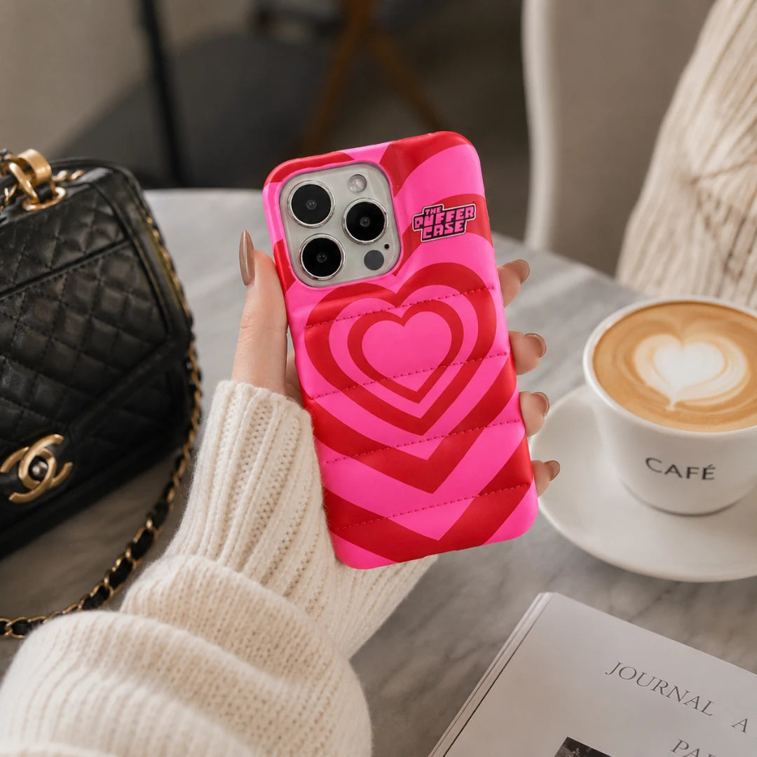 Josh & Tate - PuffHeart™ Quilted iPhone Case - Person holding a phone with a pink heart design case, next to a black handbag and a cup of coffee.