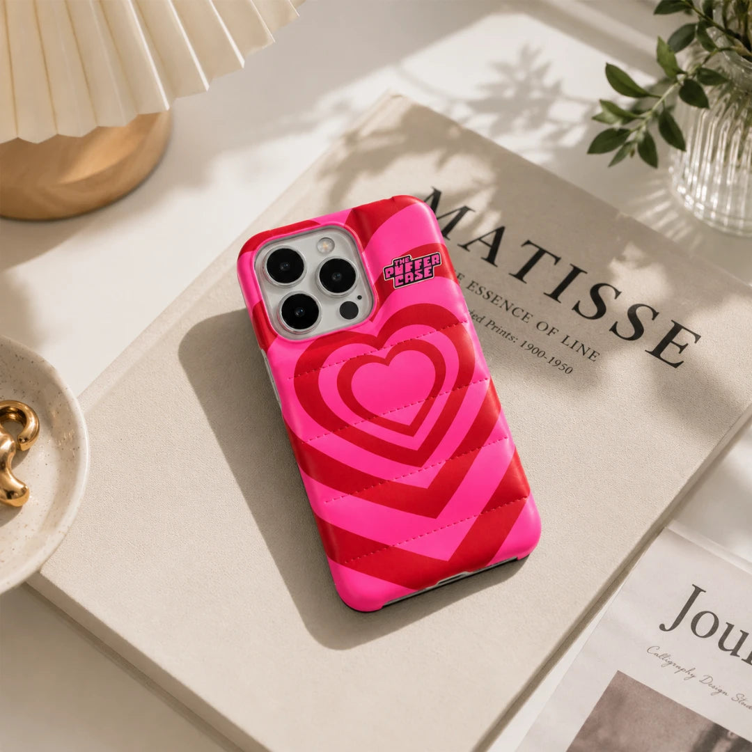 Josh & Tate - PuffHeart™ Quilted iPhone Case - Pink and red heart-patterned phone case on a surface with a magazine and plant in the background