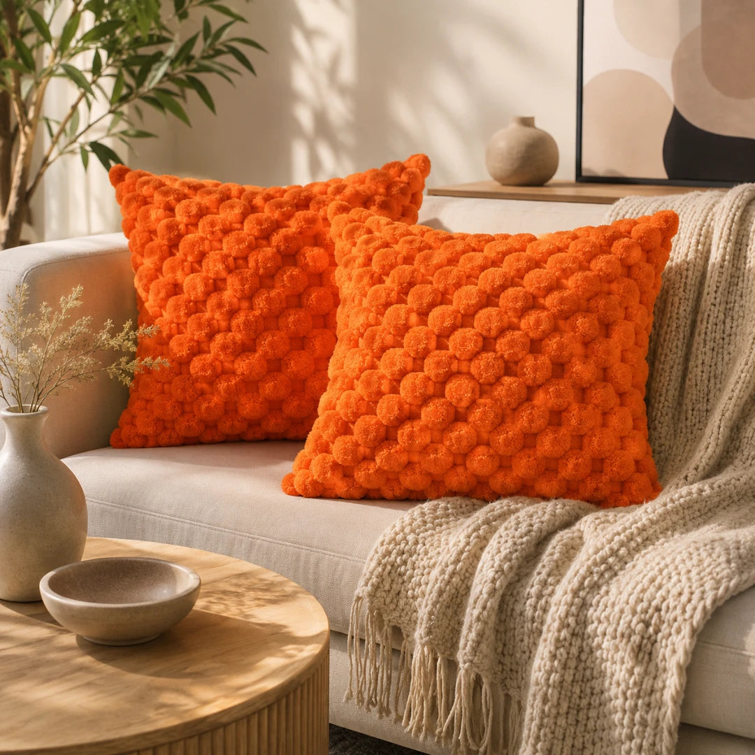 Josh & Tate - PuffKnot™ Bobble Cushion Cover - Two bright orange textured throw pillows on a light beige sofa with a cream knit blanket, styled in a warm, sunlit living room setting.