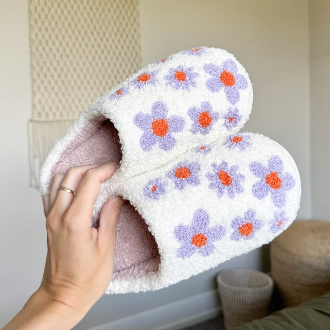 White slippers with purple flowers and orange centers held by a hand against a neutral background