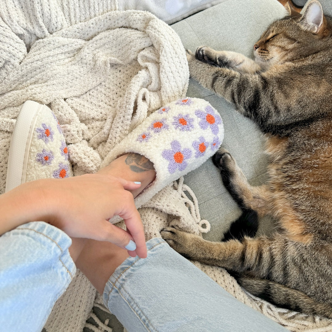 Person wearing slippers with floral design petting a cat on a cozy blanket