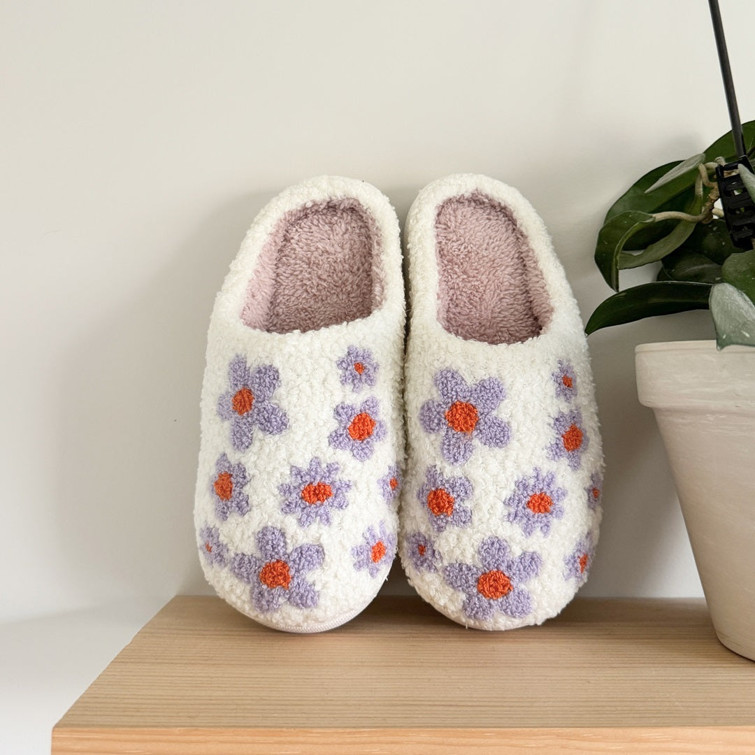 Pair of white slippers with floral patterns on a wooden surface next to a plant.