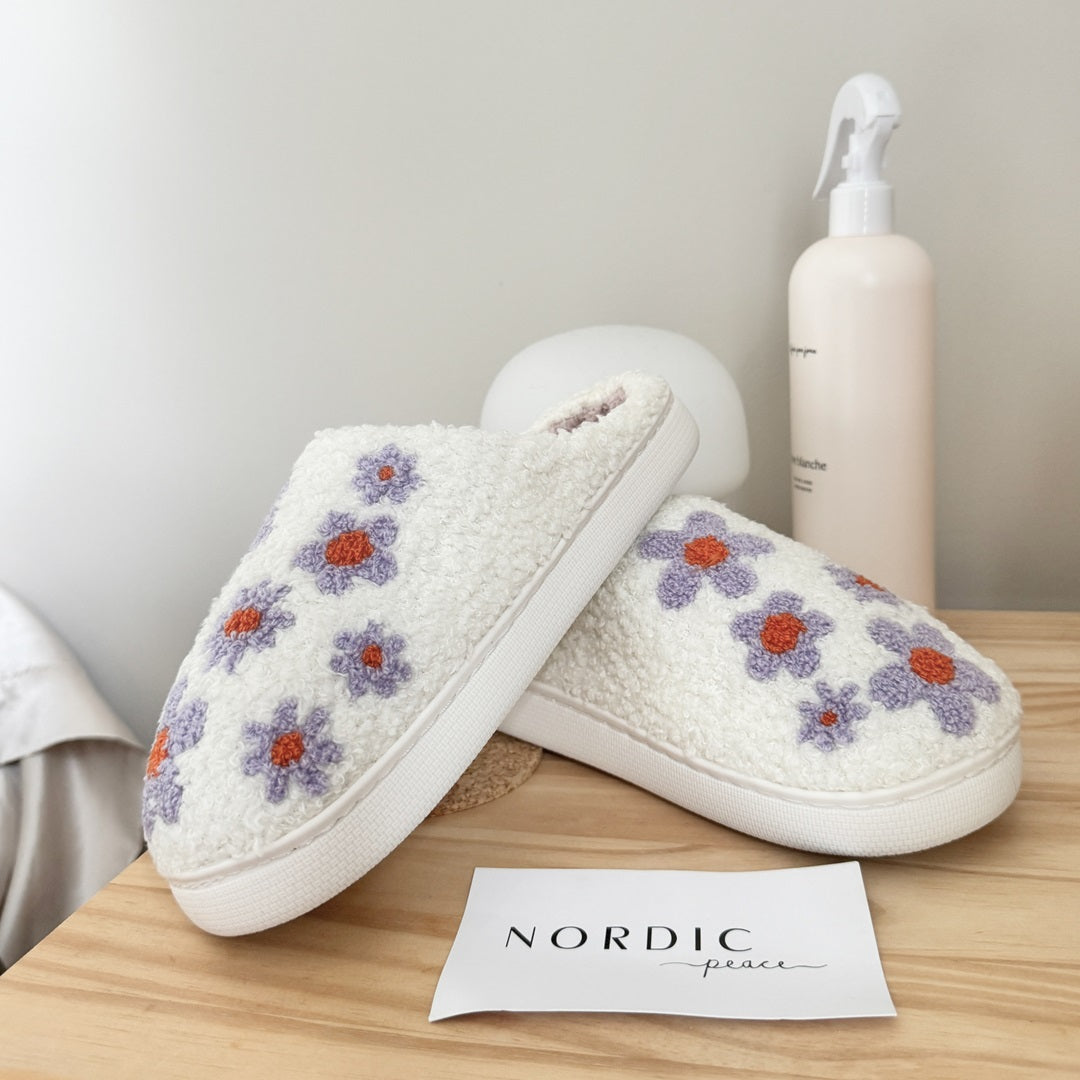 White slippers with floral patterns on a wooden surface, next to a bottle and a card with 'Nordic Peace' text.