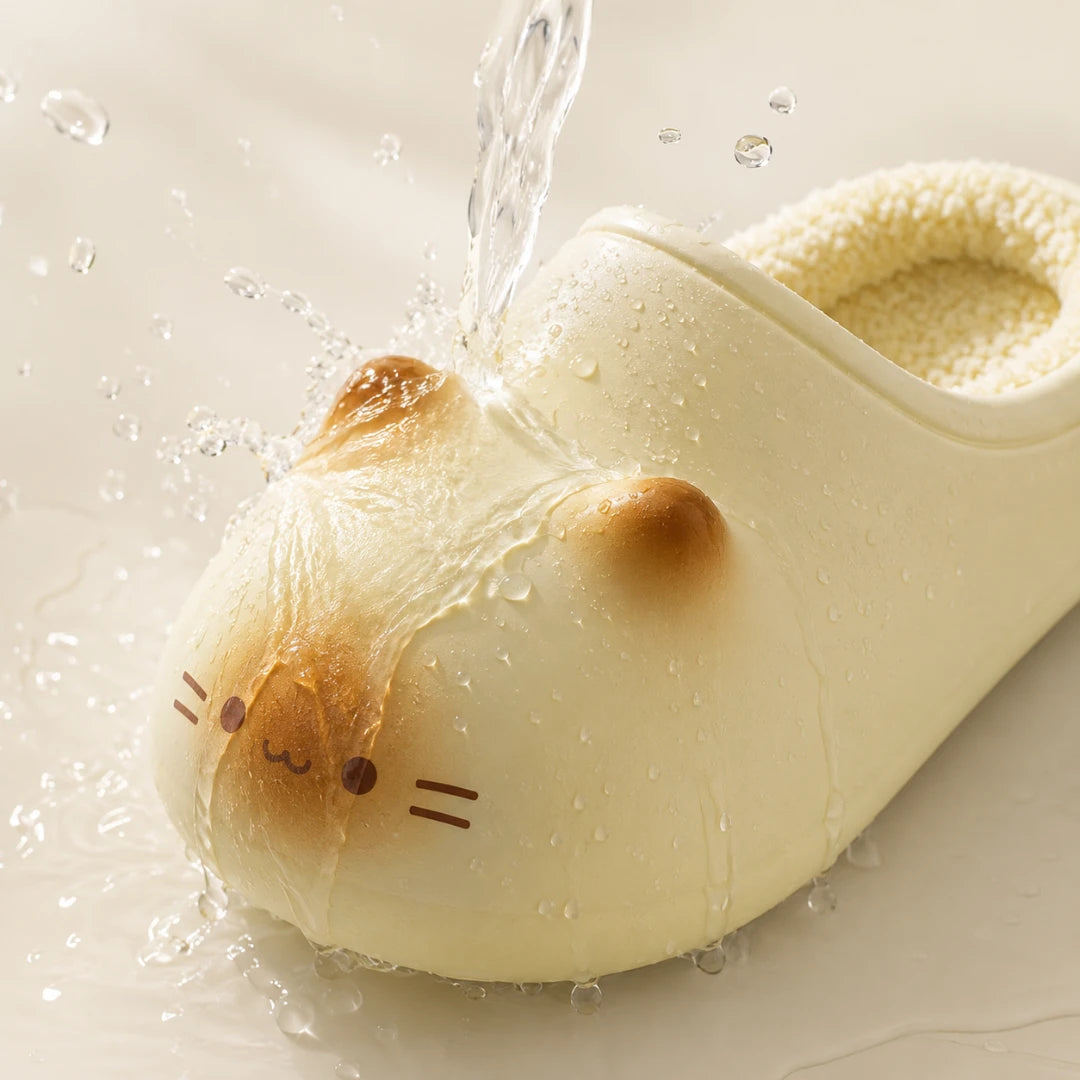 Josh & Tate - PurrPad™ Cozy Cat-Face Slippers - A close-up of a cream-colored slipper shaped like a cat face, with water being poured over it to demonstrate water resistance. The slipper has a soft, plush texture and a small brown ear accent.