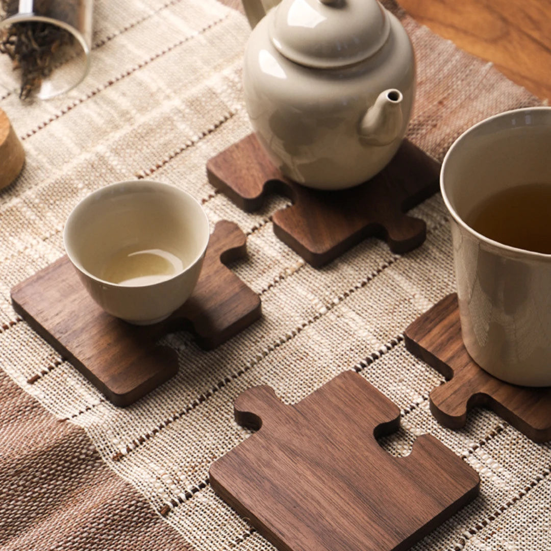 Josh & Tate - PuzzleDeck™ Wood Jigsaw Coasters - A set of puzzle-shaped dark brown wooden coasters arranged on a woven beige table runner, with a ceramic teapot, a small cup, and a larger mug partly visible.