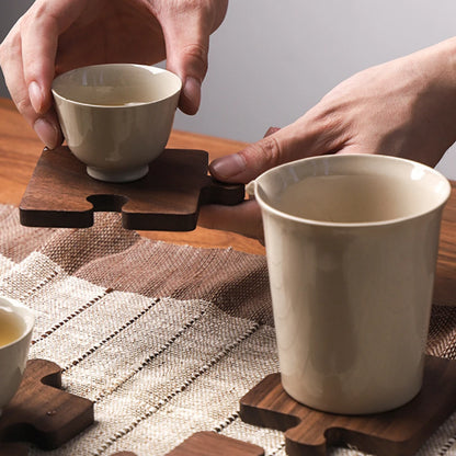 Josh & Tate - PuzzleDeck™ Wood Jigsaw Coasters - A close-up lifestyle shot of hands holding a small beige ceramic teacup on a wooden puzzle coaster, with another matching beige ceramic mug resting on a similar wooden coaster on a woven placemat.
