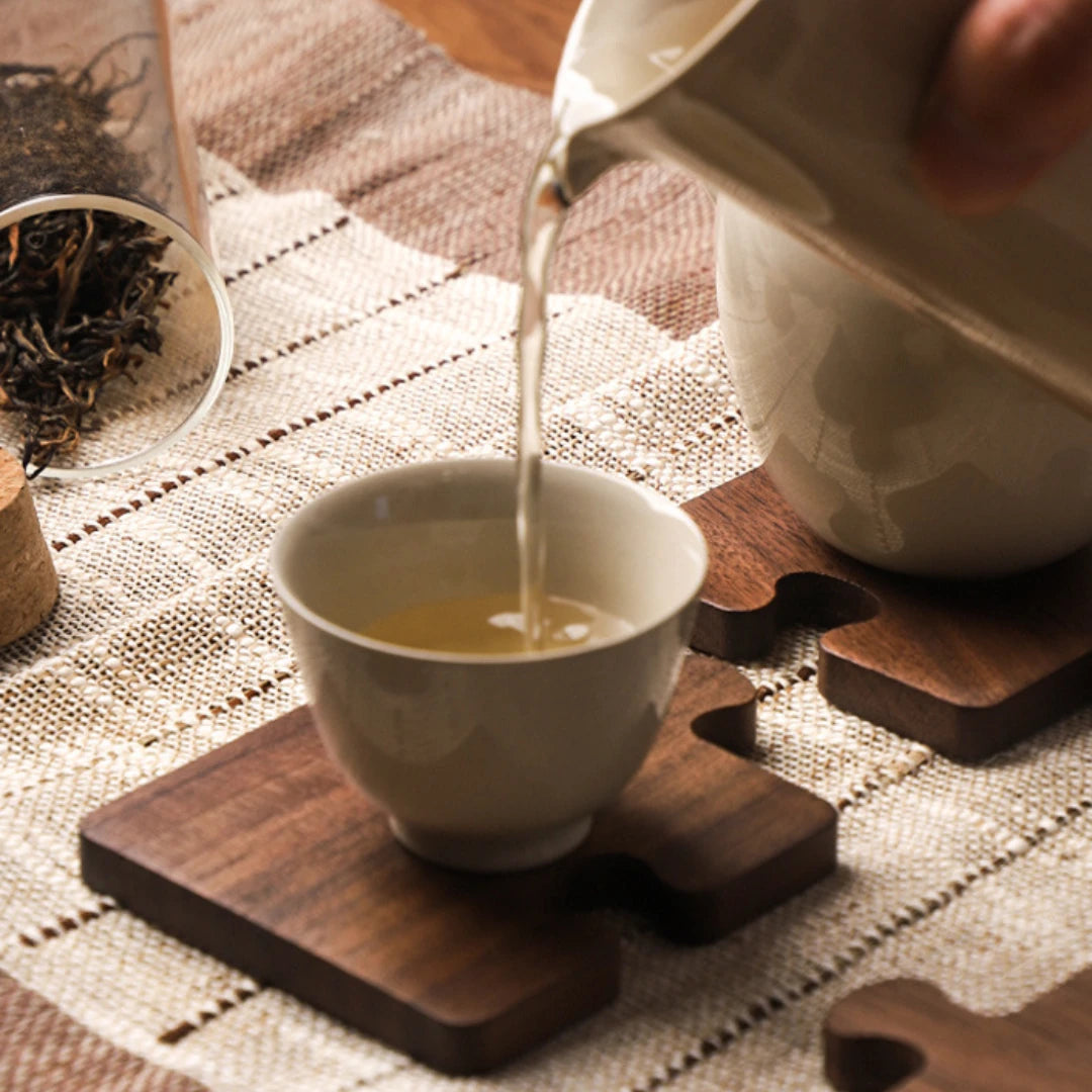 Josh & Tate - PuzzleDeck™ Wood Jigsaw Coasters - A ceramic tea cup sits on a wooden coaster while tea is being poured from a matching pitcher. A glass jar of loose-leaf tea and a woven table mat are visible in a softly lit setting.