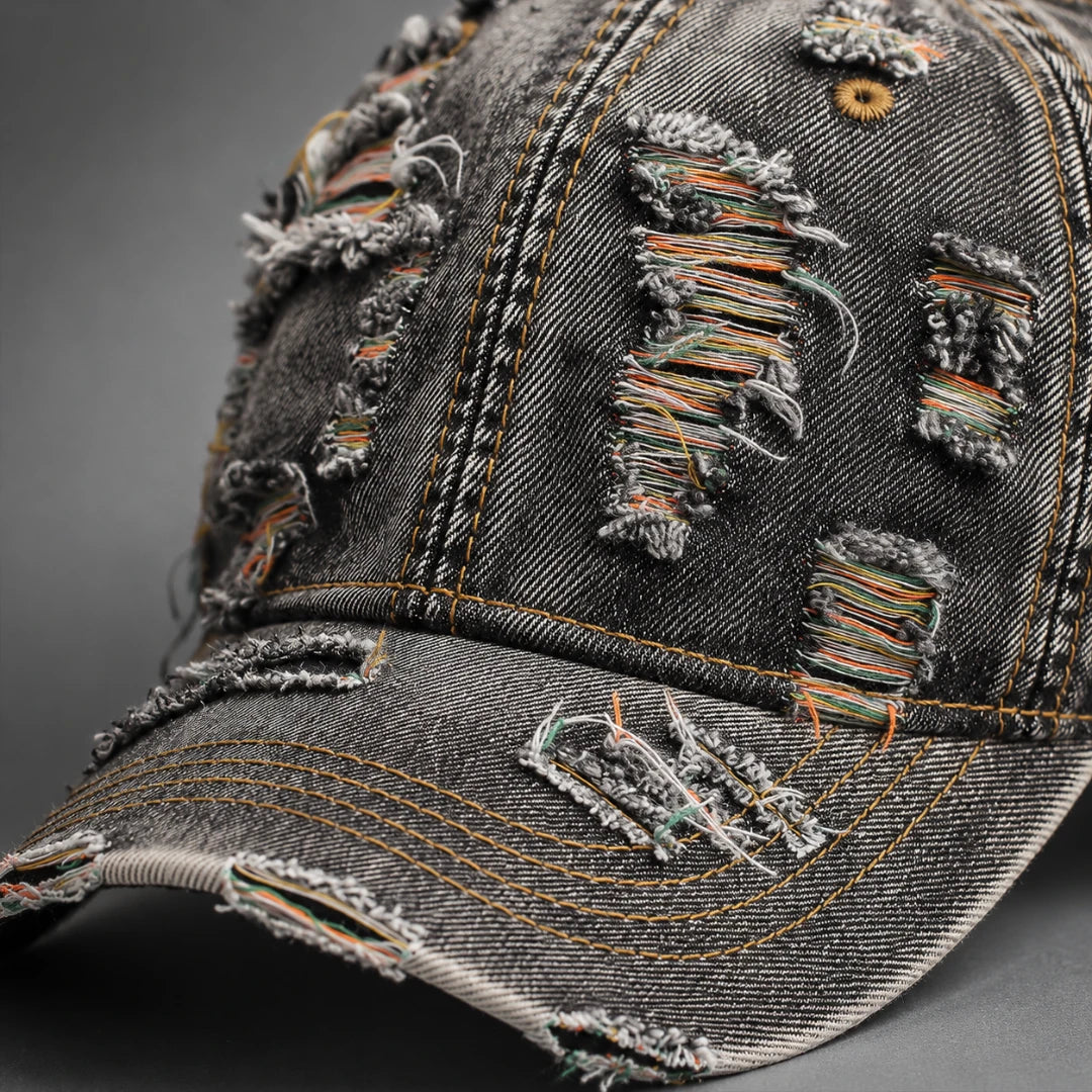 Josh & Tate - RaggedRow™ Distressed Denim Cap - A close-up of a distressed black washed denim baseball cap with ripped details showing multicolored thread fringes, on a dark gray background.