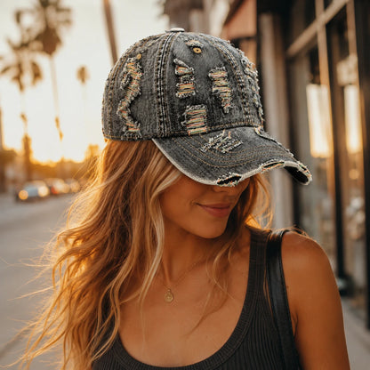 Josh & Tate - RaggedRow™ Distressed Denim Cap - A woman wearing a distressed dark gray denim baseball cap with ripped details and frayed edges, photographed outdoors on a city street at sunset.