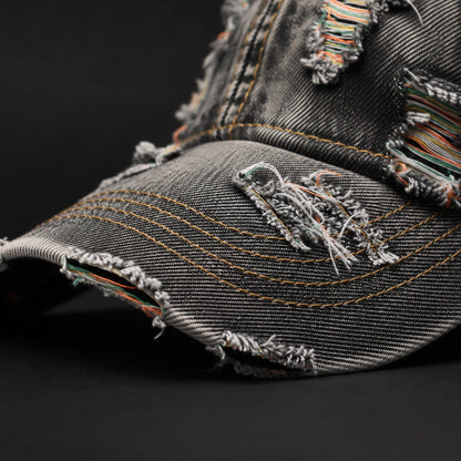 Josh & Tate - RaggedRow™ Distressed Denim Cap - Close-up of a distressed black denim baseball cap with frayed edges, multicolored thread details, and heavy contrast stitching on a dark background.