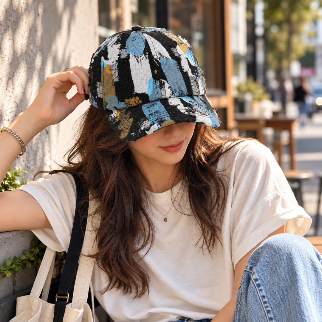 Josh & Tate - RawCanvas™ Painted Baseball Cap - A woman wearing a black baseball cap with a blue, beige, and white abstract brushstroke pattern, sitting outdoors in a sunny urban setting.