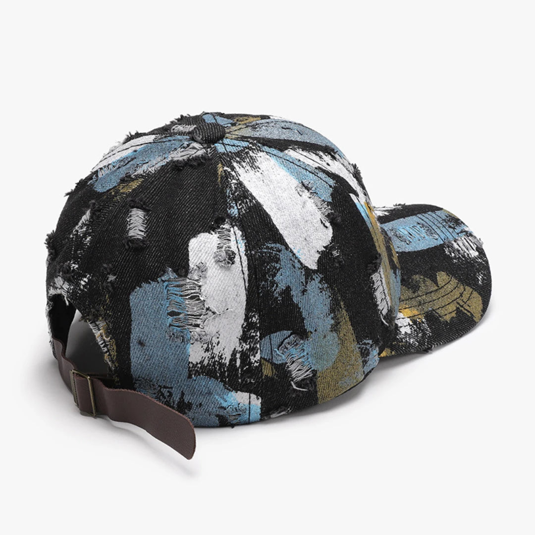 Josh & Tate - RawCanvas™ Painted Baseball Cap - A distressed baseball cap featuring an abstract paint-splatter pattern in black, light blue, white, and gold, with a brown adjustable strap.