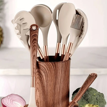 Set of kitchen utensils including spatulas and a brush in a wooden holder with vegetables in the background.