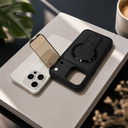 Josh & Tate - RingArmor™ MagSafe iPhone Case - A black protective phone case with a built-in kickstand and circular magnetic ring, shown resting on a light-colored book next to a smartphone. The scene is styled with a coffee cup, a green monstera leaf, and dried flowers in the background.