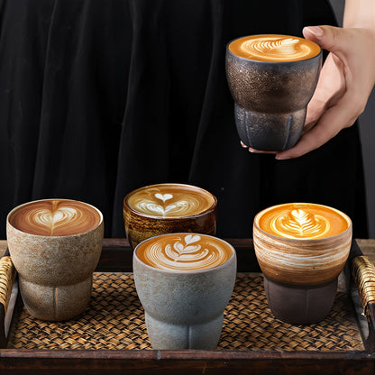 Four ceramic cups with latte art on a woven mat, one being held by a hand.